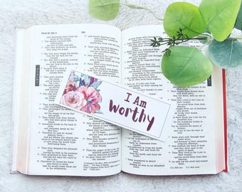 May include: A rectangular paper bookmark featuring a floral illustration with pink and blue flowers on the left side and the dark purple text "I Am Worthy" printed on the right side, resting on the open pages of a printed book.