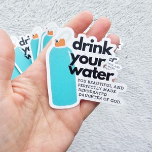Könnte beinhalten: Eine Sammlung von Aufklebern mit einem hellblauen Wasserflaschendesign mit einem orangefarbenen Deckel. Die Aufkleber tragen den Text "drink your water" und eine motivierende Botschaft. Die Aufkleber werden in einer Hand gehalten.