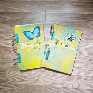 May include: Two handmade journals with yellow covers, adorned with blue butterfly illustrations and teal accents. Each journal features the text "I AM TRANSFORMED" on a torn paper strip, bound with metal rings.