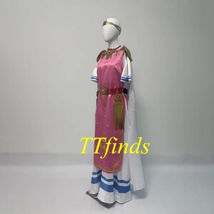 Pode incluir: Um vestido rosa e branco com detalhes dourados e uma capa branca. O vestido tem um cinto dourado com uma borla e uma bainha listrada azul e branca. O vestido est&aacute; em um manequim.