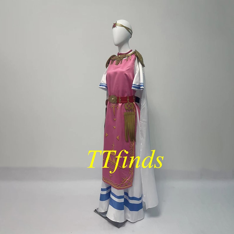 Pode incluir: Um vestido rosa e branco com detalhes dourados e uma capa branca. O vestido tem um cinto dourado com uma borla e uma bainha listrada azul e branca. O vestido est&aacute; em um manequim.