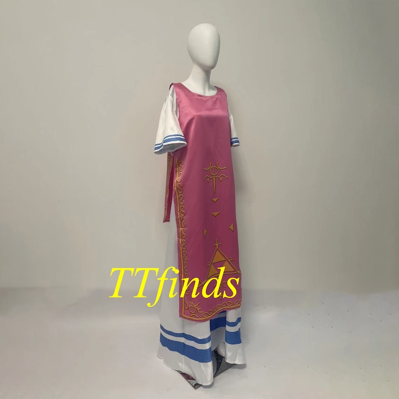 Pode incluir: Um vestido rosa e branco com um design dourado. O vestido tem uma saia longa e um corpete sem mangas. O design do vestido &eacute; um tri&acirc;ngulo estilizado com um s&iacute;mbolo solar dentro. O vestido est&aacute; em um manequim branco.