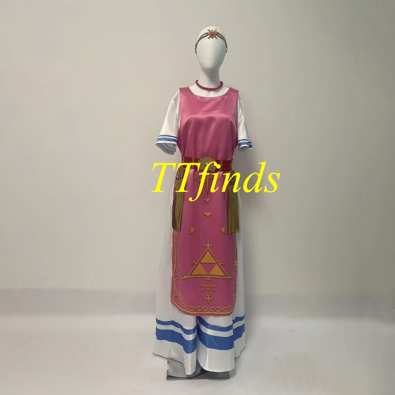 Pode incluir: Um vestido rosa e branco com um cinto dourado e um s&iacute;mbolo triangular na frente. O vestido tem uma saia branca com listras azuis e uma saia rosa com uma borda dourada. O vestido est&aacute; em um manequim branco. O texto "TTfinds" &eacute; vis&iacute;vel na imagem.