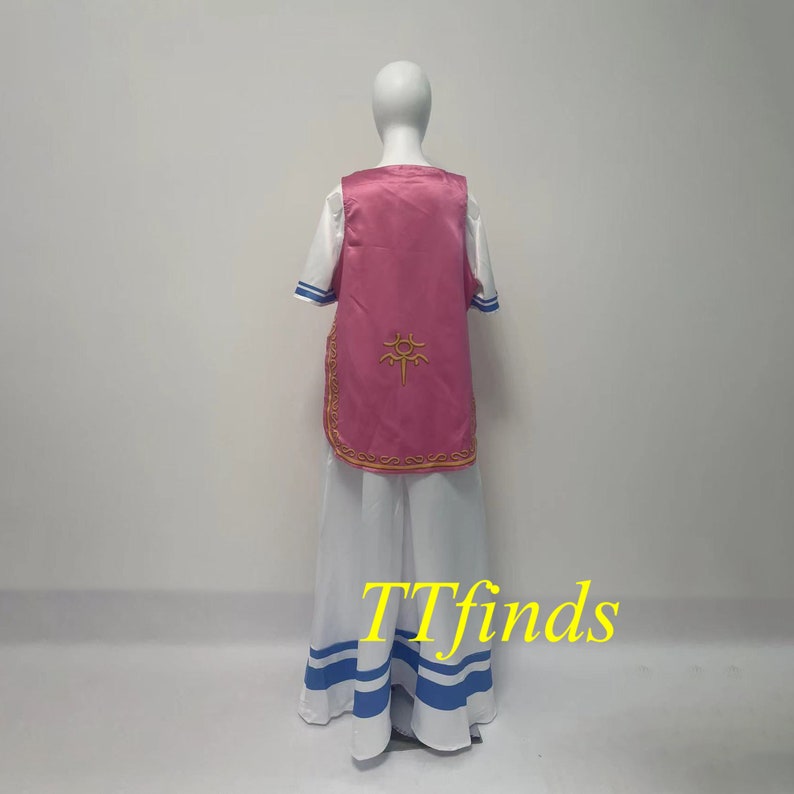 Pode incluir: Um traje rosa e branco com um s&iacute;mbolo dourado nas costas. O traje inclui uma saia longa com listras azuis, uma camisa branca com detalhes azuis e um colete rosa.