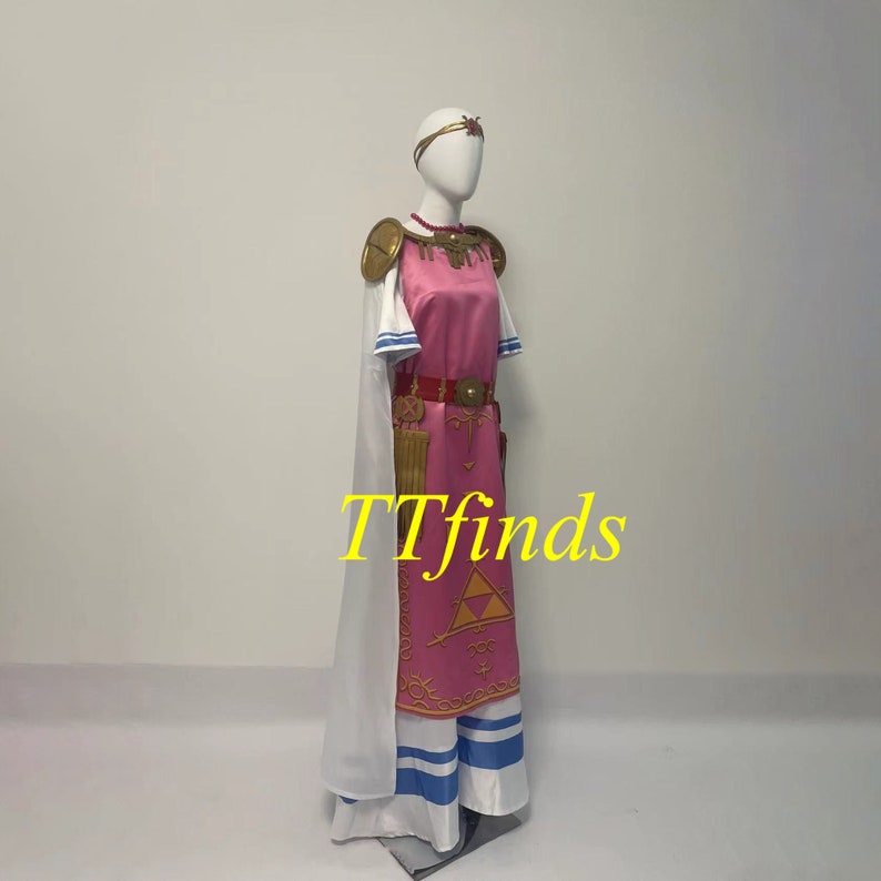 Pode incluir: Um traje rosa e branco com um cinto dourado e uma capa branca. O traje apresenta um s&iacute;mbolo triangular dourado com um olho estilizado dentro. O texto "TTfinds" &eacute; vis&iacute;vel na imagem.