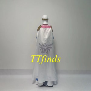 Pode incluir: Fantasia de cosplay branca com detalhes azuis e dourados. A fantasia inclui uma capa longa com um grande s&iacute;mbolo Triforce cinza nas costas. O texto "TTfinds" est&aacute; impresso em amarelo na capa.
