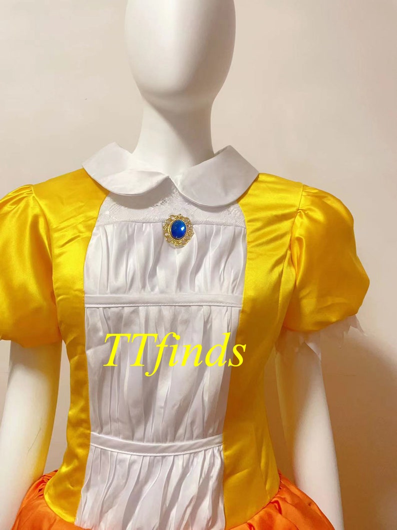 May include: A yellow and orange dress with a white bodice and a blue gem brooch. The dress has a Peter Pan collar and short sleeves. The text "TTfinds" is visible on the dress.
