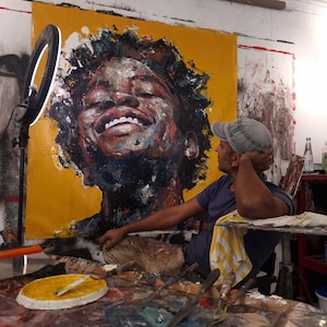 Puede incluir: Primer plano de una gran pintura de un rostro con un fondo amarillo brillante. La persona sonríe y tiene el pelo oscuro y rizado. El artista está sentado frente a la pintura, mirándola.