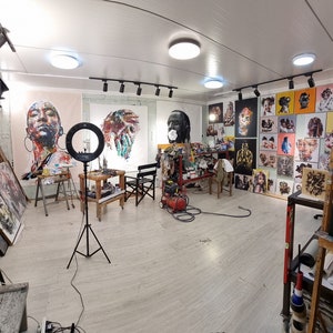 Może przedstawiać: Jasno oświetlone studio artystyczne z r&oacute;żnymi obrazami na ścianach i sztalugach. Obrazy to gł&oacute;wnie portrety os&oacute;b w kolorowych, abstrakcyjnych stylach. W pomieszczeniu znajdują się r&oacute;wnież r&oacute;żne artykuły i sprzęt artystyczny, w tym laptop, statyw i pistolet natryskowy.