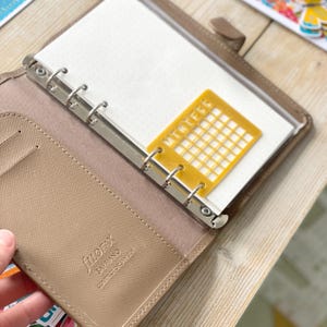 Op de afbeelding: Een bruine lederen gebonden planner met een zilveren ringband. De planner is open op een blanco pagina met een gele plastic kalenderrooster overlay.