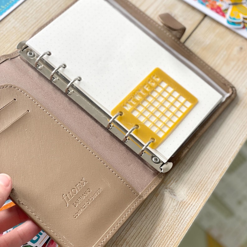 Op de afbeelding: Een bruine lederen gebonden planner met een zilveren ringband. De planner is open op een blanco pagina met een gele plastic kalenderrooster overlay.