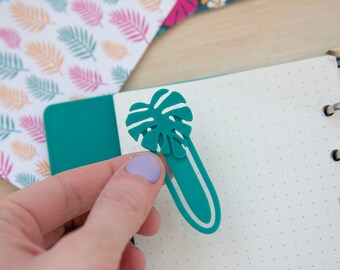 May include: Teal green leaf-shaped bookmark with a monstera leaf design. The bookmark is attached to a teal green binder.