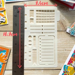 Może przedstawiać: Biała plastikowa szablon z siatką, liniami, sercami, gwiazdami i k&oacute;łkami. Szablon ma 9,4 cm szerokości i 16 cm wysokości.