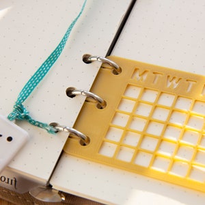 Op de afbeelding: Een gele plastic kalendergrid met de dagen van de week gelabeld "MTWTFSS" is bevestigd aan een witte planner met een turquoise lint en een witte keramische gamecontroller-hanger.