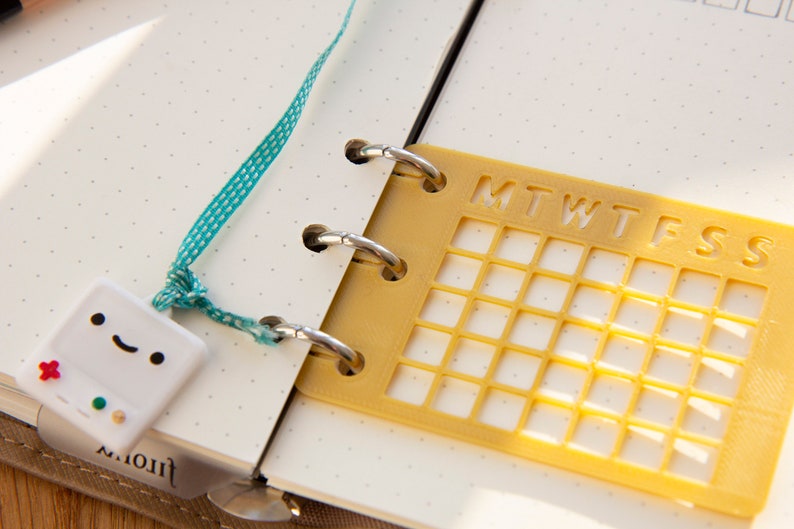 Op de afbeelding: Een gele plastic kalendergrid met de dagen van de week gelabeld "MTWTFSS" is bevestigd aan een witte planner met een turquoise lint en een witte keramische gamecontroller-hanger.