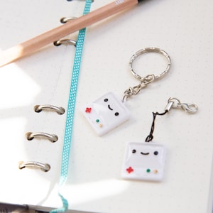 May include: Two white keychain charms shaped like retro video game consoles with happy faces and colorful buttons. The charms are attached to silver key rings.