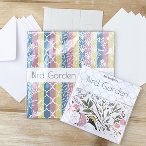 May include: A set of craft supplies featuring a pack of patterned paper and a pack of die-cut stickers. The paper pack has a variety of colorful patterns with birds, flowers, and geometric designs. The sticker pack has a blue background with white and green die-cut stickers of birds, flowers, and leaves. The paper pack is labeled "Bird Garden" and the sticker pack is labeled "Sticker Kitten Bird Garden Die-Cut Ephemera 44pcs".