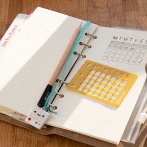 Op de afbeelding: Een bruine lederen planner met een gele plastic sjabloon voor een weekkalender en een potlood. De planner heeft een stickertabblad met de tekst "Sticker Notes".
