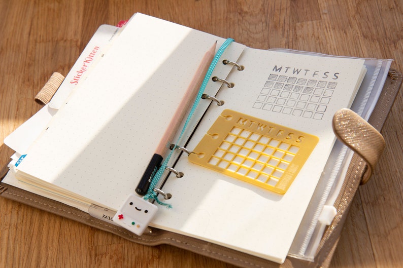 Op de afbeelding: Een bruine lederen planner met een gele plastic sjabloon voor een weekkalender en een potlood. De planner heeft een stickertabblad met de tekst "Sticker Notes".