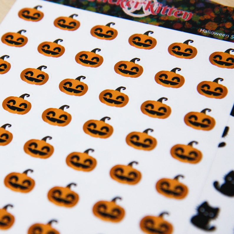 Peut inclure: Une feuille d'autocollants d'Halloween avec plusieurs visages de citrouille orange avec des yeux et des bouches noirs. Les autocollants sont sur un fond blanc avec le texte "Sticker Kitten" et "Halloween Stickers" imprim&eacute; sur la feuille.