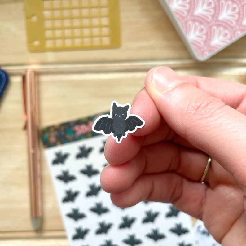 May include: A sheet of black bat stickers with a single sticker being held by a hand. The bat sticker has a smiling face.