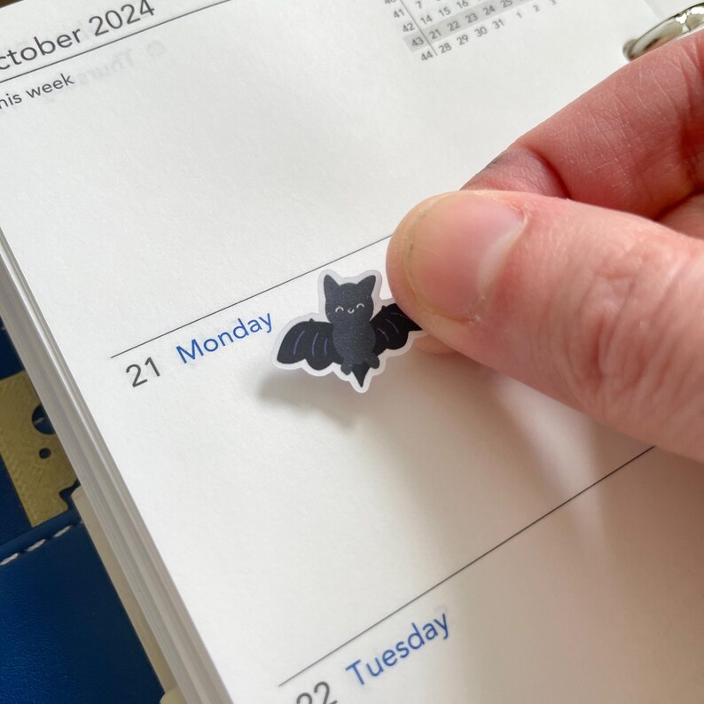 May include: A black bat sticker with a smiling face, placed on a white page of a calendar with the date "21 Monday" written in blue ink.