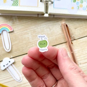 Puede incluir: Una pegatina de manzana verde con una cara sonriente y el texto "Week 15" arriba y "Apple" abajo.
