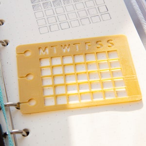 Op de afbeelding: Een geel plastic rooster met de letters MTWTFSS erop gedrukt. Het rooster is bedoeld om te gebruiken als planner of tracker.