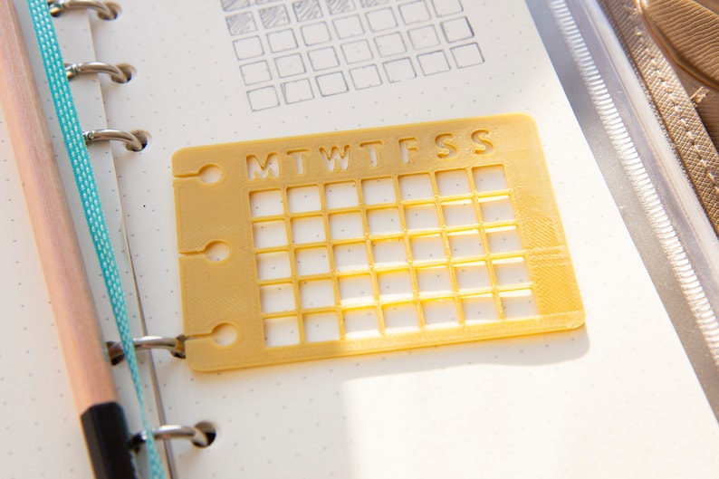 Op de afbeelding: Een geel plastic rooster met de letters MTWTFSS erop gedrukt. Het rooster is bedoeld om te gebruiken als planner of tracker.