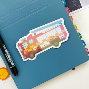 May include: A red, yellow and orange fire engine sticker with a holographic finish. The sticker is on a blue journal with a textured cover.
