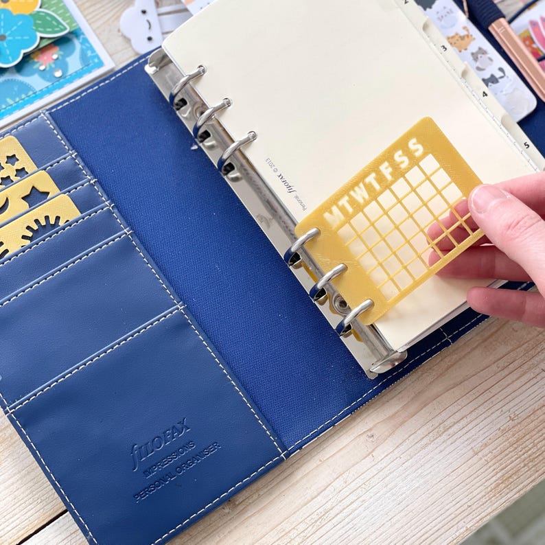 Op de afbeelding: Een blauwe Filofax persoonlijke organizer met een gouden kalenderrooster sticker bevestigd aan de ringband. De sticker heeft de dagen van de week afgekort als M, T, W, T, F, S, S.