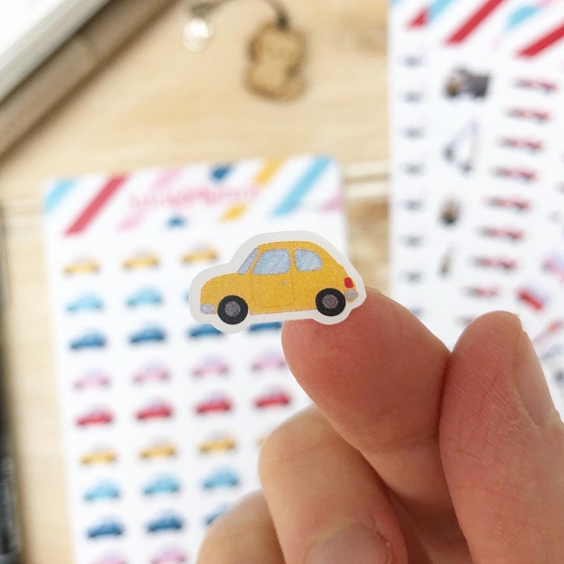 May include: A yellow car sticker with a white interior and black wheels. The sticker is on a finger.