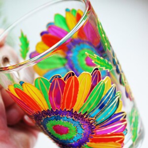 May include: A clear glass with a vibrant, hand-painted floral design. The artwork features a large, colorful sunflower with petals in shades of red, orange, yellow, green, and purple, outlined in gold. The glass is held by a hand.