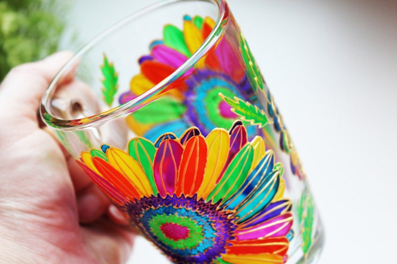 May include: A clear glass with a vibrant, hand-painted floral design. The artwork features a large, colorful sunflower with petals in shades of red, orange, yellow, green, and purple, outlined in gold. The glass is held by a hand.