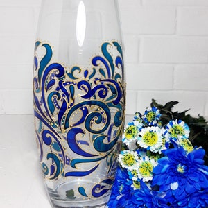 May include: A clear glass vase with a hand-painted design in shades of blue and gold. The vase features an intricate, swirling pattern. A bouquet of blue and white flowers sits beside the vase, adding a touch of color.