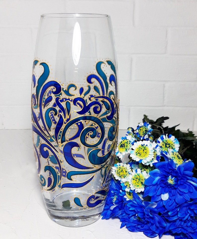 May include: A clear glass vase with a hand-painted design in shades of blue and gold. The vase features an intricate, swirling pattern. A bouquet of blue and white flowers sits beside the vase, adding a touch of color.