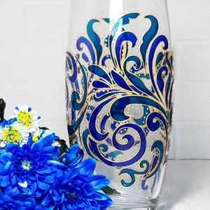 May include: A clear glass vase with a hand-painted design. The design features swirling patterns in shades of blue and teal, accented with gold details. The vase is cylindrical and stands upright. Blue flowers are in the foreground.