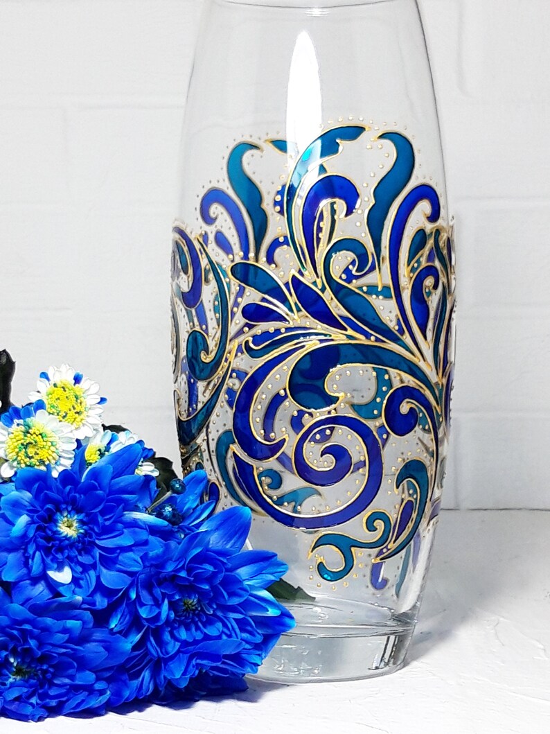 May include: A clear glass vase with a hand-painted design. The design features swirling patterns in shades of blue and teal, accented with gold details. The vase is cylindrical and stands upright. Blue flowers are in the foreground.