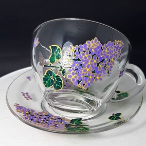 Może przedstawiać: Przezroczysty szklany zestaw filiżanki i spodka do herbaty. Filiżanka i spodek zdobione są kwiatowym wzorem z fioletowymi kwiatami bzu, złotymi akcentami i zielonymi liśćmi. Filiżanka ma uchwyt.