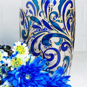 May include: A clear glass vase with a hand-painted design in shades of blue and gold. The vase features a swirling, floral pattern. A bouquet of blue and white flowers is in front of the vase.