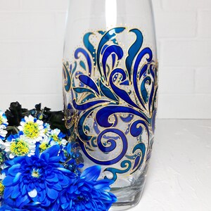 May include: A clear glass vase with a hand-painted design in shades of blue and gold. The vase features an intricate, swirling pattern. Blue flowers are arranged in front of the vase, adding a pop of color.
