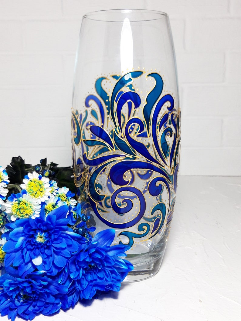 May include: A clear glass vase with a hand-painted design in shades of blue and gold. The vase features an intricate, swirling pattern. Blue flowers are arranged in front of the vase, adding a pop of color.