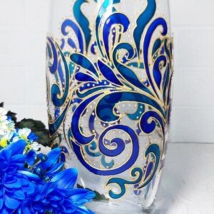 May include: A clear glass vase with a hand-painted design. The design features swirling blue and teal patterns with gold accents. The vase is cylindrical and has a wide opening, suitable for displaying flowers.