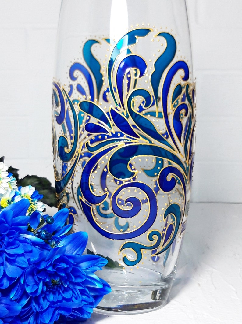 May include: A clear glass vase with a hand-painted design. The design features swirling blue and teal patterns with gold accents. The vase is cylindrical and has a wide opening, suitable for displaying flowers.