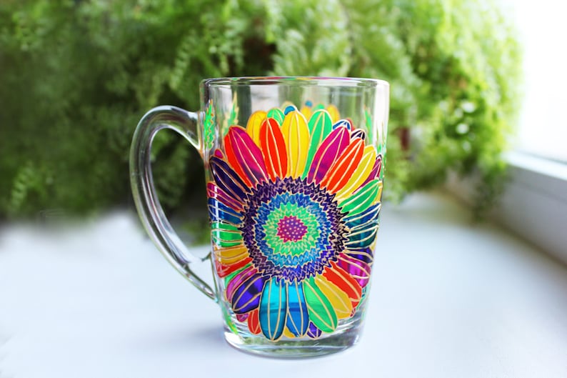 May include: Clear glass mug with a vibrant, hand-painted sunflower design. The flower features a spectrum of colors, including red, orange, yellow, green, blue, and purple. The mug has a clear handle and is set against a blurred green background.