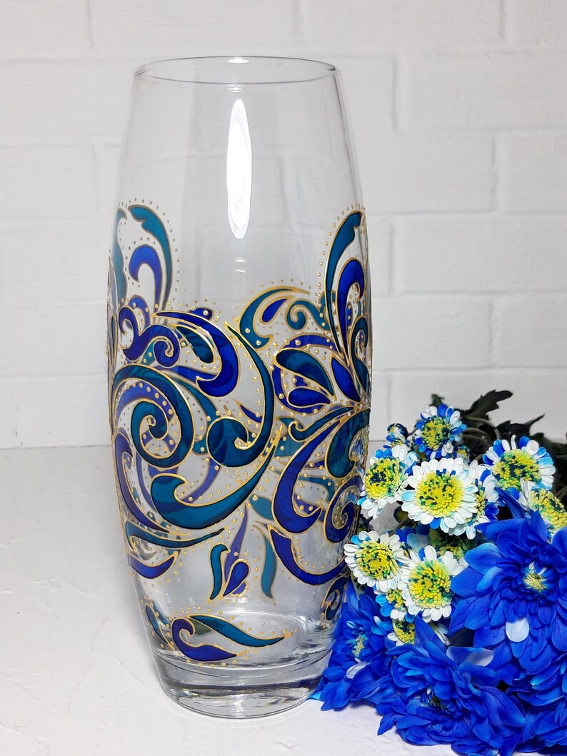 May include: A clear glass vase with a hand-painted design featuring swirling blue and teal patterns with gold accents. The vase is tall and cylindrical, with a slight flare at the top. A bouquet of blue and white flowers sits beside the vase.