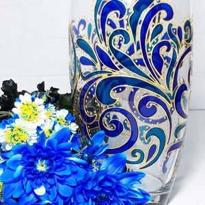 May include: A clear glass vase with a hand-painted design in shades of blue and gold. The vase features an intricate, swirling pattern. A bouquet of blue and white flowers is in front of the vase.