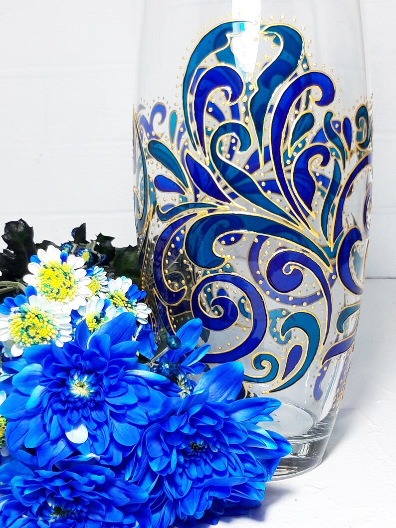 May include: A clear glass vase with a hand-painted design in shades of blue and gold. The vase features an intricate, swirling pattern. A bouquet of blue and white flowers is in front of the vase.