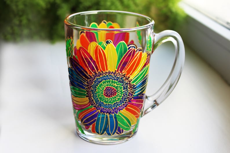 May include: Clear glass mug with a vibrant, hand-painted sunflower design. The sunflower features bright yellow, orange, red, purple, and blue petals with gold accents. The mug has a clear handle and is suitable for hot or cold beverages.