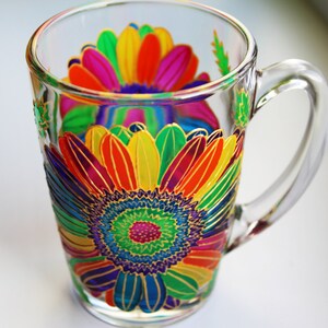 May include: Clear glass mug with a vibrant, hand-painted floral design. The colorful flower features petals in shades of red, orange, yellow, green, and purple, outlined in gold. The mug has a clear handle and is suitable for hot or cold beverages.